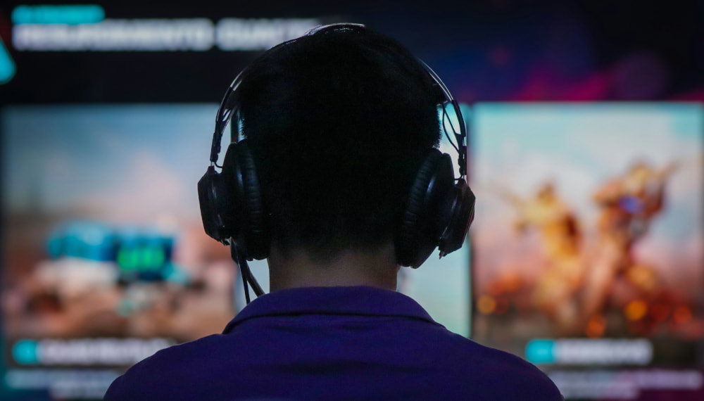 Gamer at his desk with a headset on looking at monitors where a shooter game is displayed, viewed from behind.